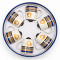 Six white ceramic espresso cups arranged in a circular presentation box, featuring a gold Medusa head logo, blue Greek key patterns, and black decorative bands.