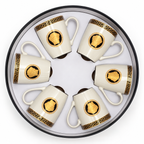 Six white ceramic coffee mugs arranged in a circle inside a round presentation box. Each mug features a gold Greek key border and a central gold Medusa head motif on a black oval background.