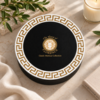 A round black gift box with a white border and gold Greek key pattern, featuring a central gold crest and the text 'The House of FLORENCE Classic Medusa Collection.' The box is styled on a light stone surface with olive branches, a candle, fabric, and a book nearby.