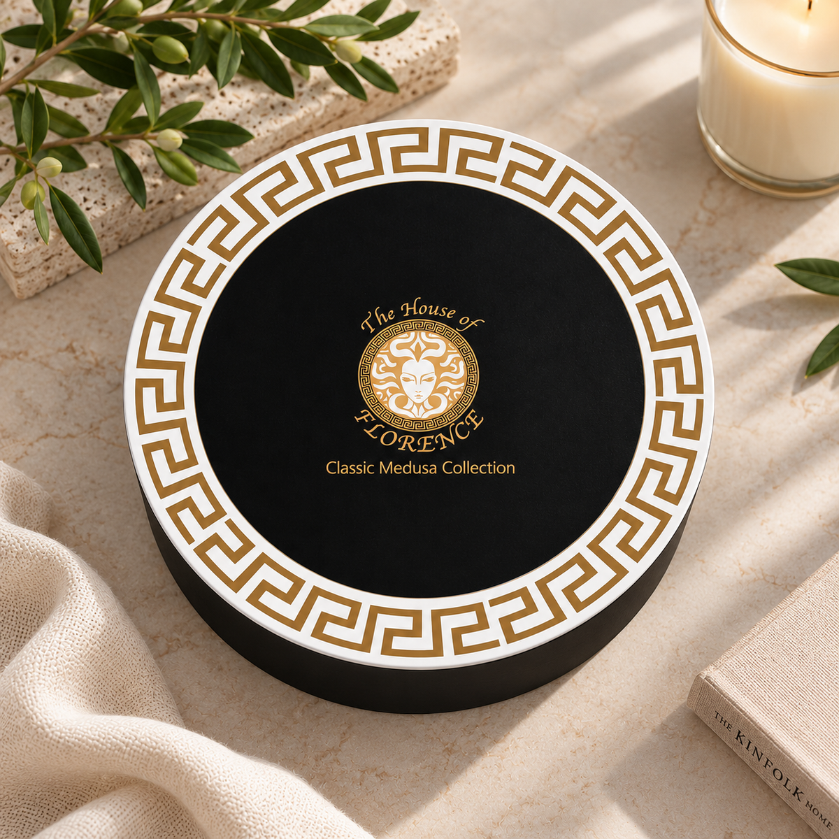 A round black gift box with a white border and gold Greek key pattern, featuring a central gold crest and the text 'The House of FLORENCE Classic Medusa Collection.' The box is styled on a light stone surface with olive branches, a candle, fabric, and a book nearby.