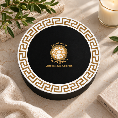 A round black gift box with a white border and gold Greek key pattern, featuring a central gold crest and the text 'The House of FLORENCE Classic Medusa Collection.' The box is styled on a light stone surface with olive branches, a candle, fabric, and a book nearby.